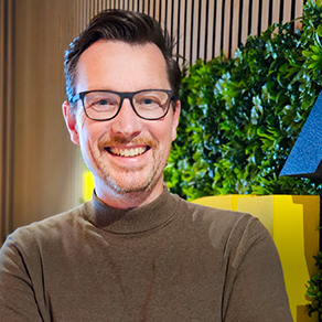 Portret van een glimlachende man met bril en bruine trui, staand voor planten en houten lamellen