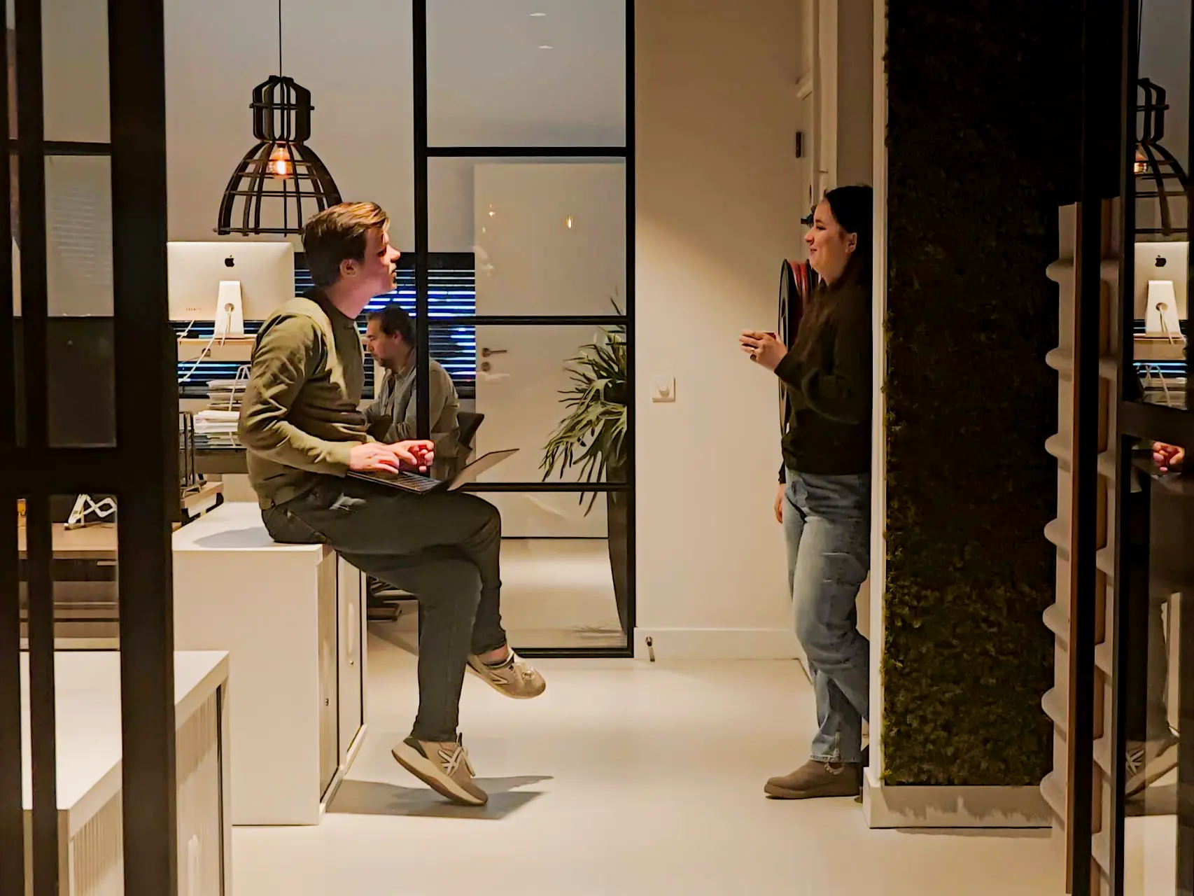 Twee mensen praten in een modern kantoor, terwijl een man op een bureau zit met een laptop