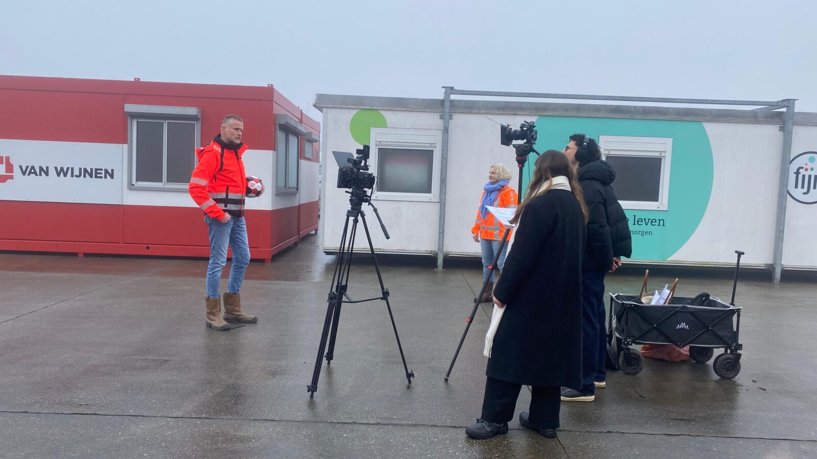 Buitenopname van een filmploeg met camera’s en een man in een oranje jas die een bal vasthoudt voor tijdelijke bouwcontainers bij van Wijnen