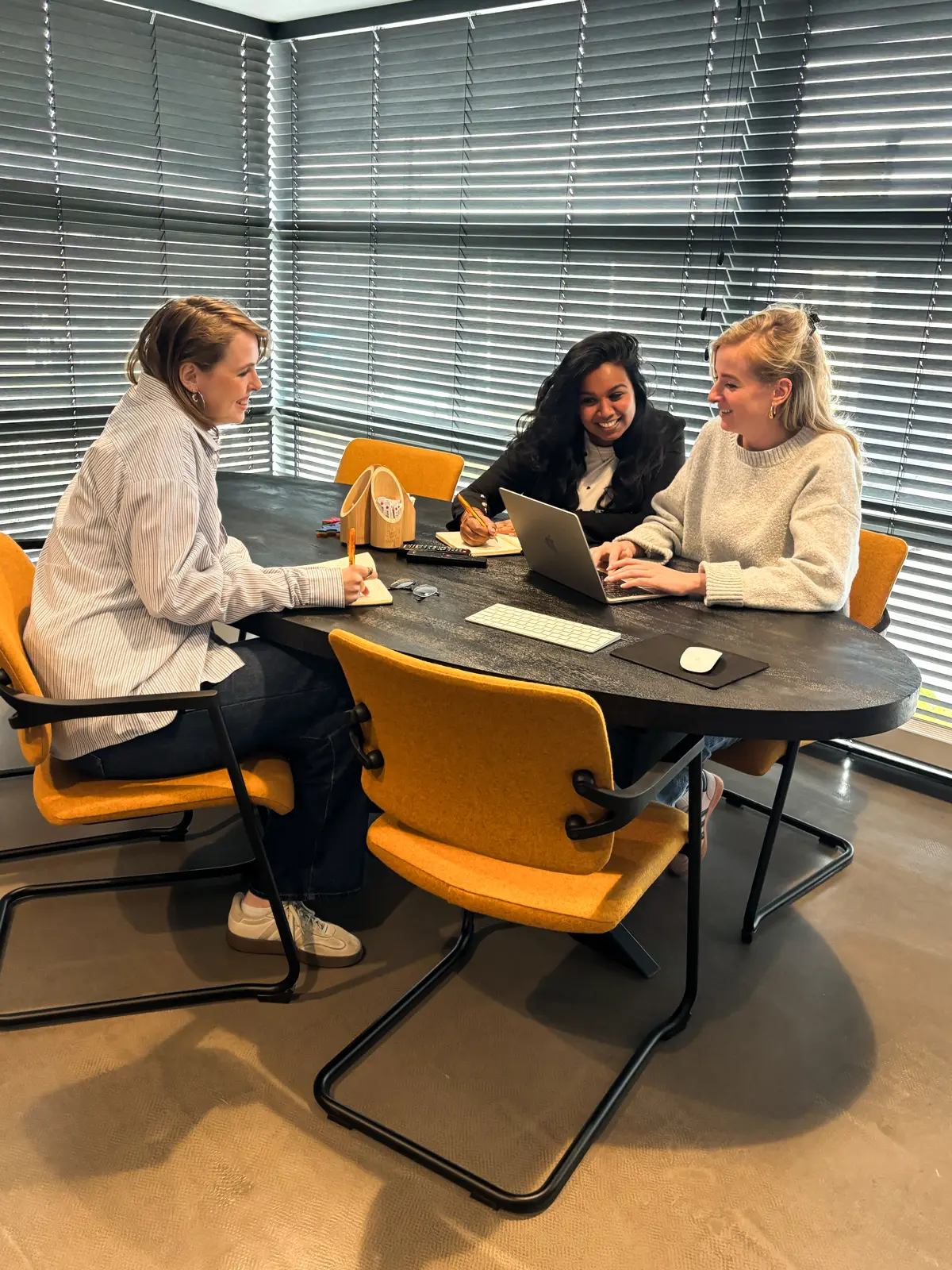 Drie vrouwen overleggen aan een tafel met laptop en notitieblokken in een vergaderruimte