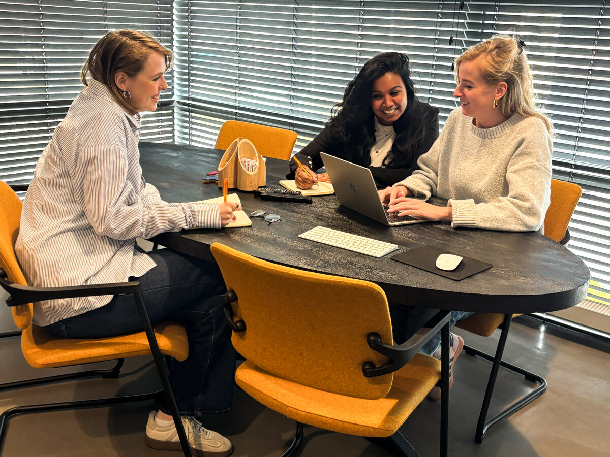 Drie vrouwen overleggen aan een tafel met laptop en notitieblokken in een vergaderruimte