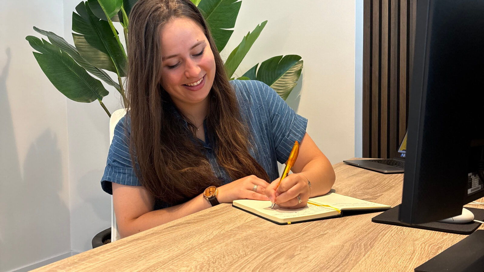 Meisje in blauwe blouse en lang donkerbruin haar achter bureau die in een schrift schrijft op kantoor